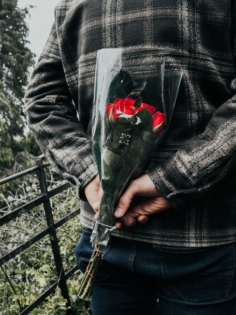 A person wearing a dark checkered jacket holding a bouquet of fresh red and white roses wrapped in clear plastic, with their hand supporting the stems. The roses are vibrant, with slightly open petals