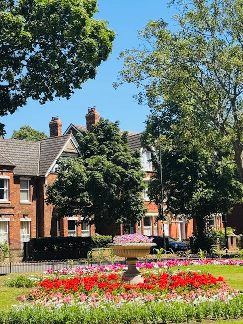 A vibrant flower bed featuring a mix of bright red, pink, and white blooms, situated in a park with lush green grass and surrounded by large, leafy trees. In the background, there are classic red-bric