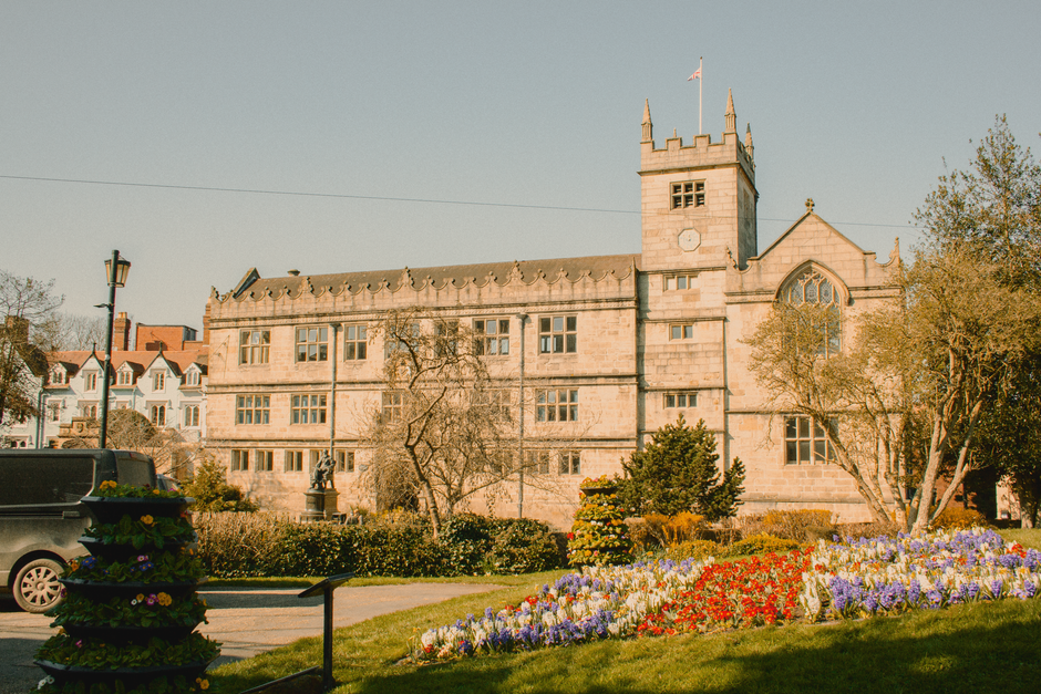 A floral arrangement featuring a bouquet of fresh flowers held by a person outside a historic stone building with a tower, surrounded by a landscaped garden with colorful tulips, hyacinths, and primro
